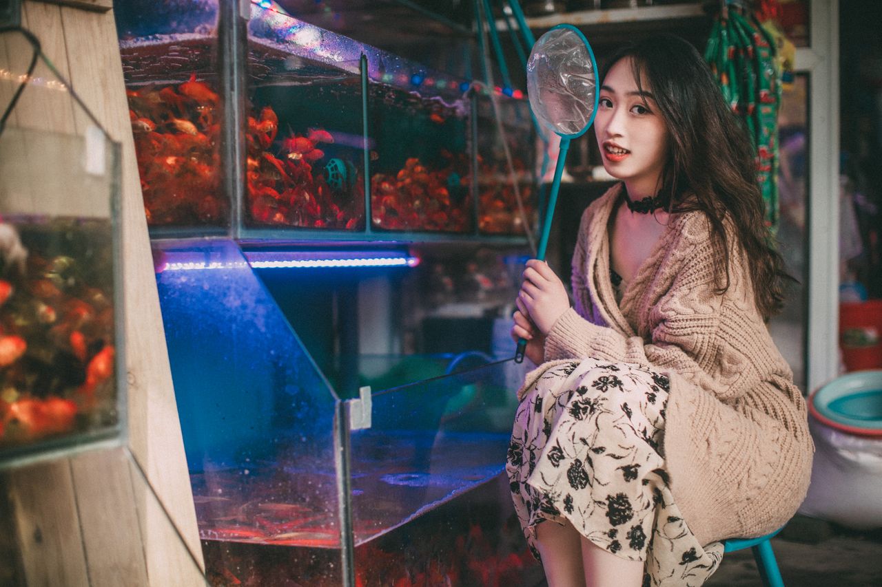 A young woman poses with a net next to vibrant fish tanks in an aquarium store, showcasing an engaging pet shop scene.